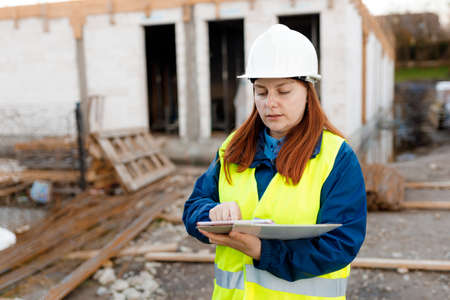 Young architect woman in white hardhat and safety vest using digital tablet outdoors. Female...