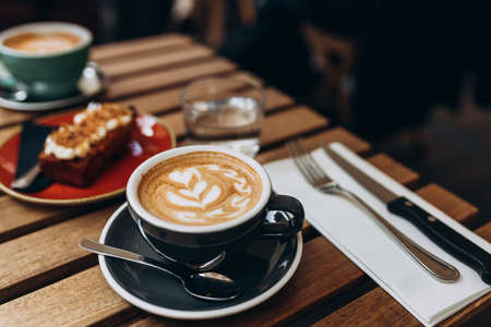 Two black Cups of cappuccino with latte art. Coffee in ceramic cup with glass of water and cakes on brown wooden table in cafe. Concept of easy breakfast. Top viewの写真素材