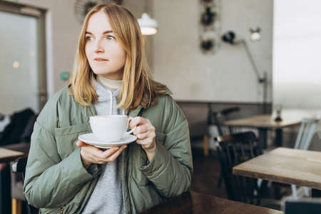 Portrait of young happy blond woman drinking coffee at table at cafe. Mature woman in a cafeteria holding hot cappuccino in ceramic white cup, Concept of easy breakfast. Mockupの写真素材