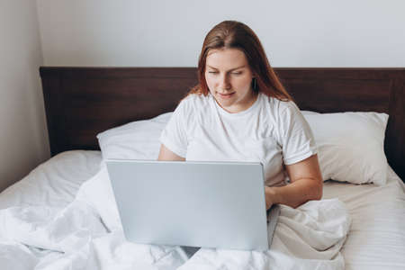 Portrait of beautiful young redhead woman with laptop on bed on a lazy day. Online education, working or shopping. Technology woman concept for alternative office freelanceの写真素材