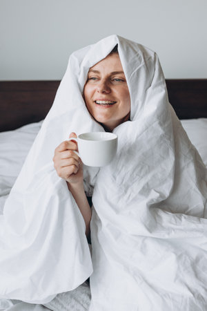 Young happy redhead woman with cup of coffee sitting in bed under blanket at home bedroom. comfort or home weeekend concept, mockup of mugの写真素材