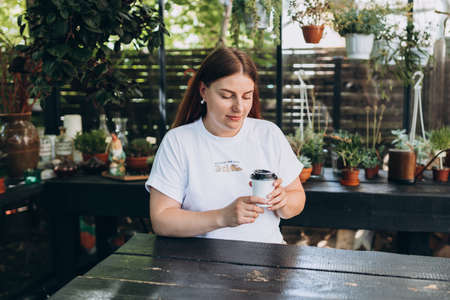 Young beautiful woman with paper white cup of coffee sitting at summer rustic cafe. Mockup. Female...