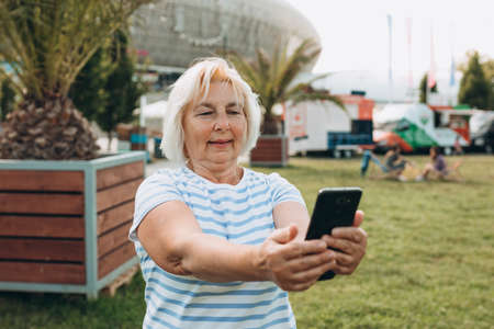 Mature blonde woman in casual summer clothes browsing smart phone while streets of city. Caucasian...