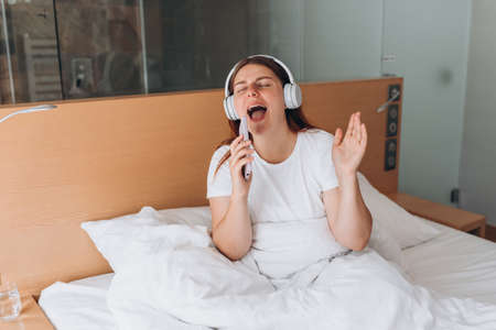 Closeup portrait of a lovely young girl listening to music through wireless earphones and singing on bed at home. Music lover enjoying music. Woman record voice by mobie cell phone dictaphoneの写真素材