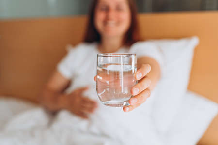 Pregnant woman with glass of water sitting on bed at home. Millennial Caucasian woman wake up in...
