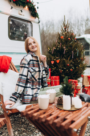 Blond woman warming with Cup of Coffee at Campsite. Cheerful young girl standing near modern caravan Trailer outdoors. New Year and Christmas concept.の写真素材