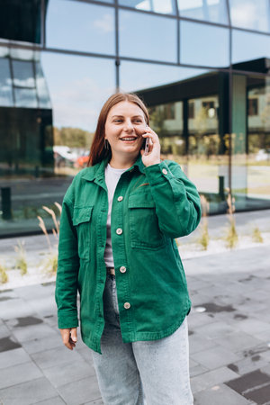 Beautiful woman in trendy wear spending time on street using smartphone. Happy Redhead girl talking on the mobile phone, warm sunny day. Urban lifestyle concept, technology bannerの写真素材