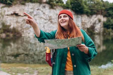 Stylish hipster woman holding paper map, wearing backpack and red hat looking at mountain view while relaxing in nature. Travel and wanderlust concept. Amazing chill moment.の写真素材