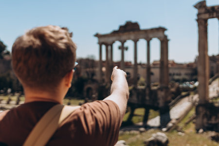 A happy man tourist is standing near the Roman Forum, old ruins at the center of Rome, Italy. Historical imperial Foro Romano from panoramic point of view. Men pointing finger, sunny dayの写真素材