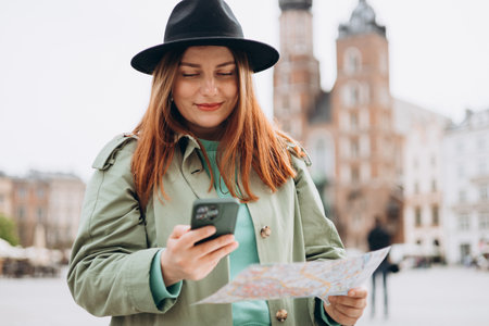 Beautiful stylish woman in hat walking on Market Square in Krakow on autumn day and holding mobile phone and map. Phone Communication. Urban lifestyle concept. Check social networks, booking hotelの写真素材