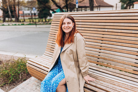 Happy cheerful young redhead woman looking at camera and sitiing on the bench in the park on city street, Urban lifestyle concept. Traveler, rest timeの写真素材