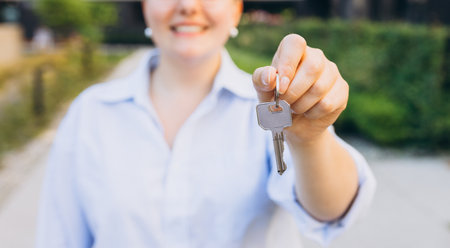 Attractive young woman showing key while standing outdoor against new house. Real estate agent holding key on street backgroundの写真素材