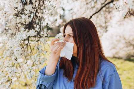 Sneezing young redhead woman with nose wiper among blooming trees in park. Portrait of sick women sneezes in white tissue, suffers from rhinitis and running nose. Symptoms of cold or allergy.の写真素材