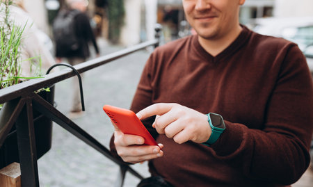 Millennial male browsing website on modern cellphone device using 4g wireless at street terrace. Handsome and relaxed man sitting at a cafe table outdoorsの写真素材