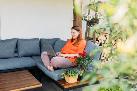 Cheerful happy young redhead woman relaxing with laptop in the yard of the house in summer. Casual beautiful woman is using computer sitting on a sofa at homeの写真素材