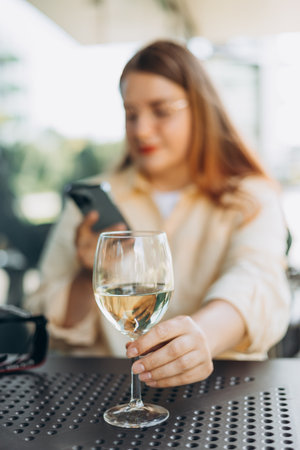 White wine glass in an outdoor cafe. Happy Woman holding glass with wine and using mobile phoneの写真素材