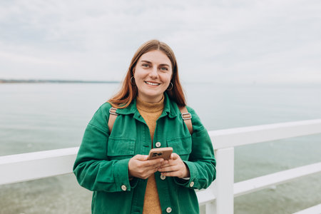 Beautiful young smiling woman with backpack using smartphone during walk at the beach. Hipster girl...