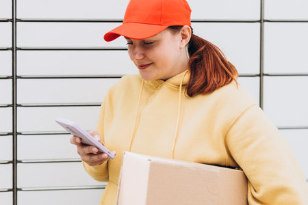 Young woman in red hat picks up parcel from automatic post office machine, Courier standing with phone and small box. Concept of fast delivery to automatic self lockersの写真素材