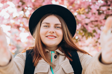 Young happy woman wearing hat taking selfie on spring city street. 30s girl smiling enjoying spring...