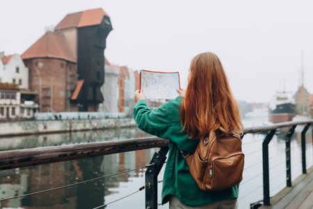 Attractive young female tourist is exploring new city. Redhead girl with backpack holding a paper...