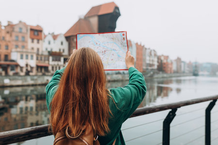 Attractive young female tourist is exploring new city. Redhead girl with backpack holding a paper...