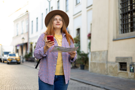 Picture of pretty young woman staying on the street holding phone and map in hands. 30s tourist...