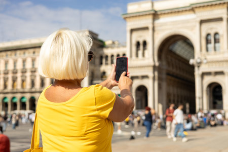 Beautiful senior blonde woman taking selfie on Duomo square, Galleria Vittorio Emanuele II shopping arcade in Milan, Italy. Traveling Europe in summer. Attractive 60s female is exploring cityの写真素材
