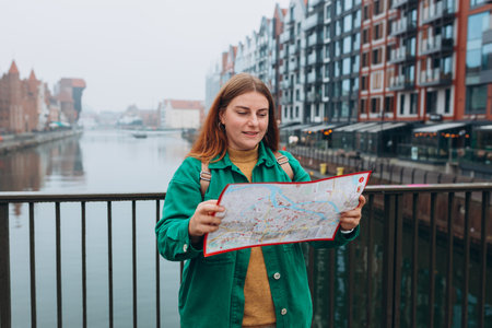 Attractive young female tourist is exploring new city. Redhead girl with backpack holding a paper map on city street in Gdansk. Traveling Europe in autumn. Famous Zuraw crane, Motlawa riverの写真素材