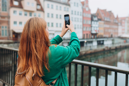 Back view of young European tourist makes photo or video on smartphone at city of Gdansk, Poland. Hipster girl with backpack hold on smart phone gadget outdoors, back viewの写真素材