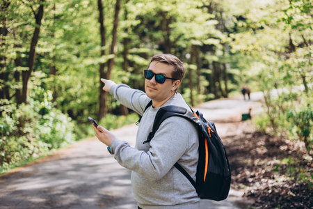 Happy men with phone walking on city park and checks smartphone. Blonde male in sport clothes with backpack pointing finger outdoors, he hiking on woodlandの写真素材