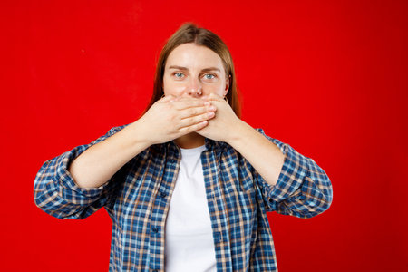 Young 30s woman covering closed mouth with hands open eyes. Speak no evil concept isolated red...
