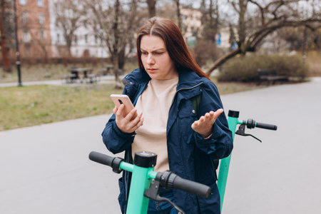 Portrait of young woman with confused face, holding smartphone near scooter, using mobile...