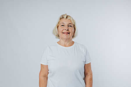Beautiful senior blonde woman smiling and looking at camera over white background. Human emotions,...