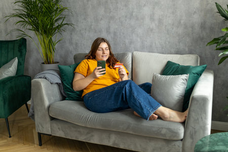 Cheerful young woman in casual clothes sit on couch spending time in living room at home. Rest relax good mood leisure lifestyle concept. 30s female Using phone, hold credit bank card, full bodyの写真素材
