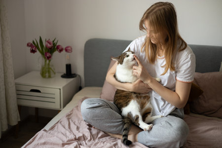 Young blonde 30s woman is resting with a Stripped White cat on the bed at home one spring day. Morning at home. Cute cat licking fingers of female owner in daytime at home. Feeding.の写真素材