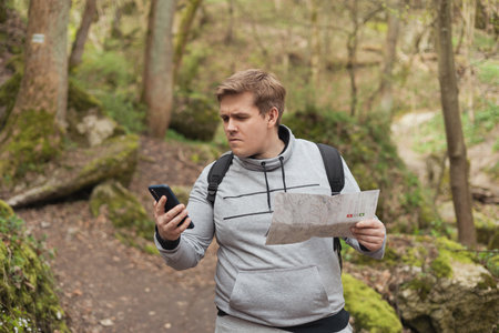 A young man tries to find a phone signal in the forest. Searching for mobile connection in wild nature on vacation. Angry man tourist hiker holding smart phone and paper mapの写真素材