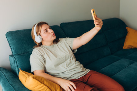 Happy young female taking a selfie photo with kiss at home. 30s woman with headphones recording self video, talking on online call and relaxing on a sofaの写真素材