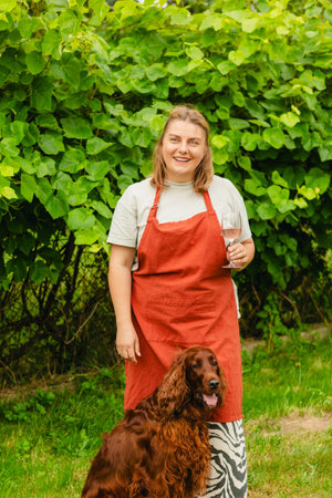 30s Woman in apron with dog tasting pink wine in vineyard. Portrait of pretty young woman holding...