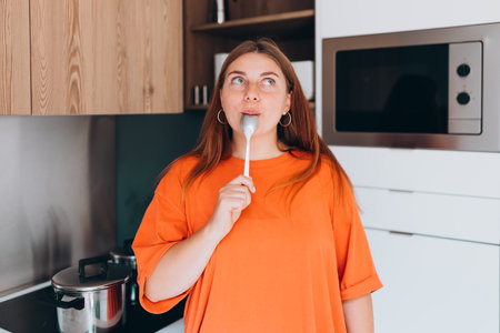 Emotional woman eating tasty meal at the kitchen, girl standing near stove at home. Domestic life and leisure moments. High quality photo. Portrait of excited hungry young woman holding spoon in mouthの写真素材