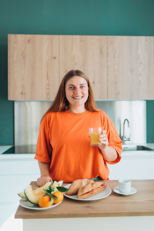A happy 30s woman standing in a kitchen, holding a glass of fresh orange juice at home. Domestic life and leisure moments. Healthy diet. Young woman with glass of drink at table in kitchen.の写真素材