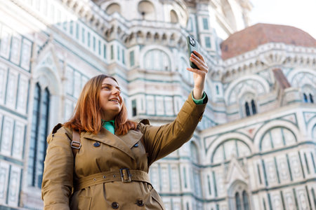 Happy redhead Girl making selfie photo on urban background. Use technology concept, Traveling Europe in autumn. 30s woman using smartphone near famous Duomo cathedral in Florence.の写真素材