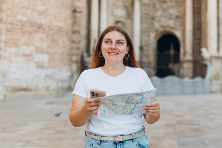 Attractive female tourist is exploring new city. Redhead woman holding a paper map near the Virgin Saint Mary, Valencia Cathedral. Traveling Europeの写真素材