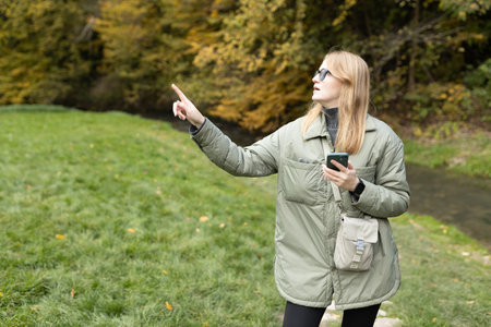 Hiker blonde woman holding smart phone and pointing finger something near river. Travel, technology and relax concept. High quality photoの写真素材