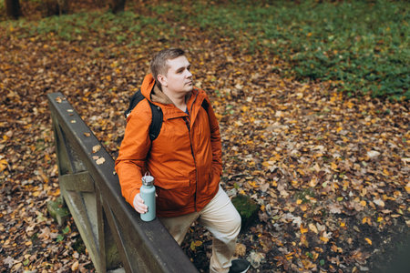 Blonde young man with thermos of tea wearing backpack standing on autumn forest trail, hiking alone. Freedom and active lifestyle concept. High quality photoの写真素材