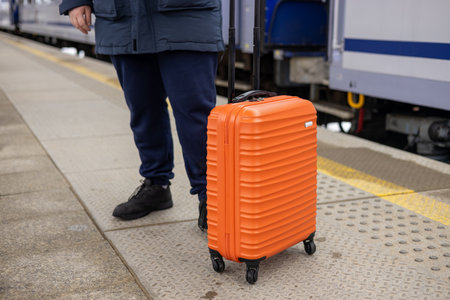 A young man with a small orange suitcase waits for a train at the train station for traveling. Lifestyle, people, adventure and tourism concept.の写真素材