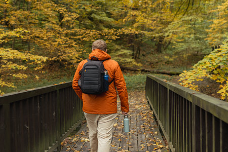 Blonde young man with thermos of tea wearing backpack standing on autumn forest trail, hiking alone....