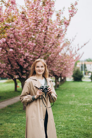 Young beautiful smiling hipster 30s woman in trendy clothes with camera. Carefree woman posing on bloom trees background. Girl in a flowering garden. Women day bannerの写真素材