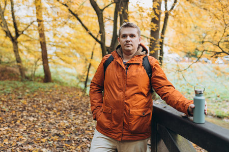 Blonde young man with thermos of tea wearing backpack standing on autumn forest trail, hiking alone....
