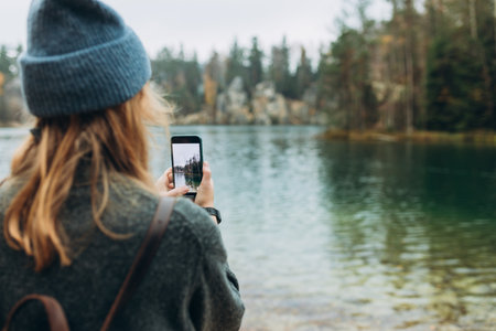 Person in hat is recording content with a smartphone near lake on wild nature background. Stylish 30s girl with phone walking outdoors. Travel, adventure. Concept of an active lifestyle.の写真素材