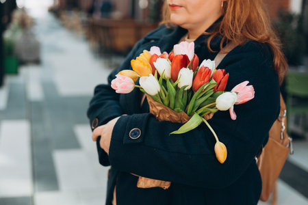 Beautiful young woman with spring tulips flowers bouquet walking at city street. Happy girl posing outdoors. Spring portrait of pretty female on urban background. High quality photo.の写真素材